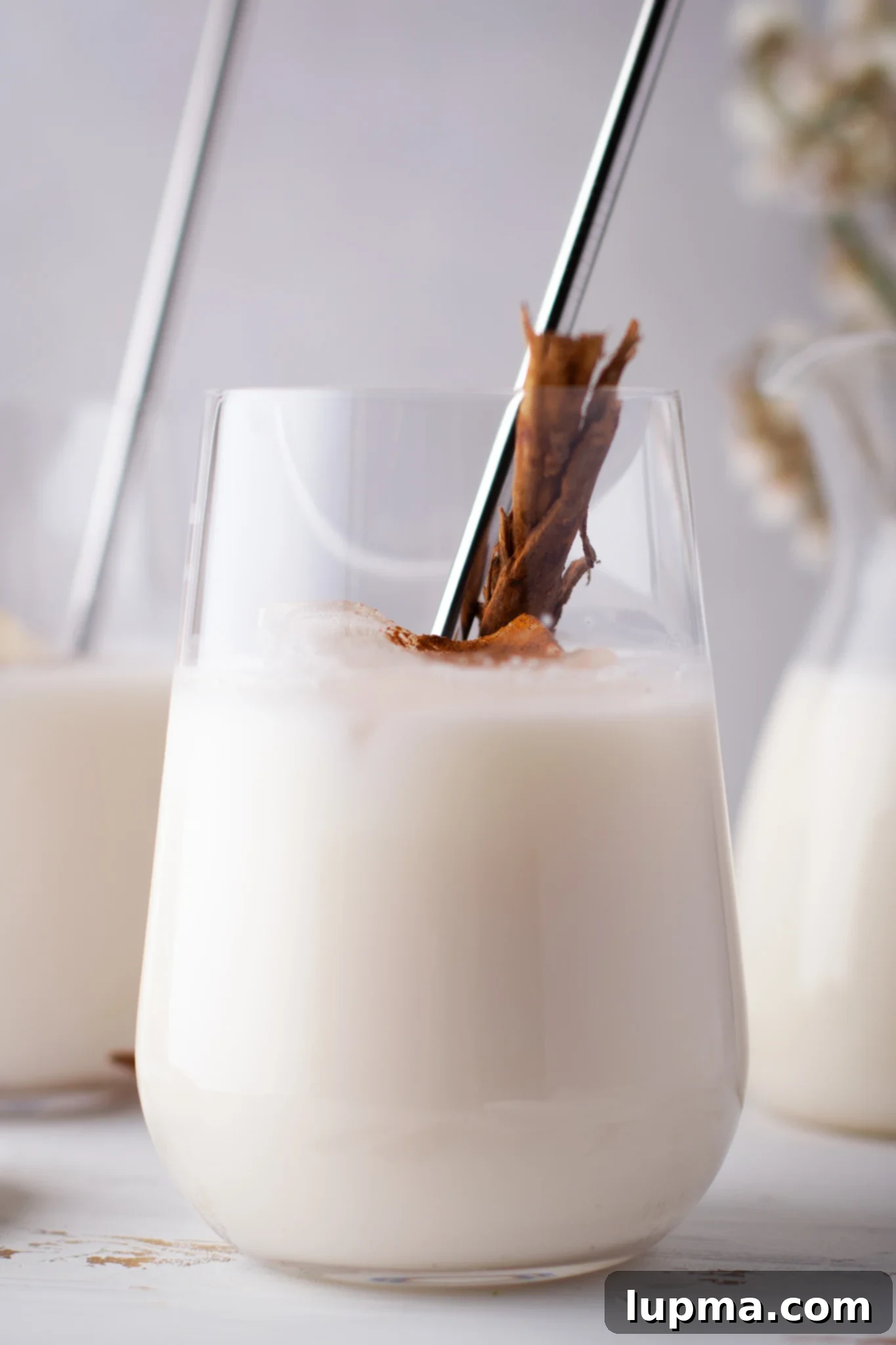 The Ultimate Horchata Recipe 2 A tall glass of homemade Horchata, garnished with a cinnamon stick and a sprinkle of ground cinnamon, resting on a rustic wooden surface. The drink is a milky white color, indicating its creamy texture.