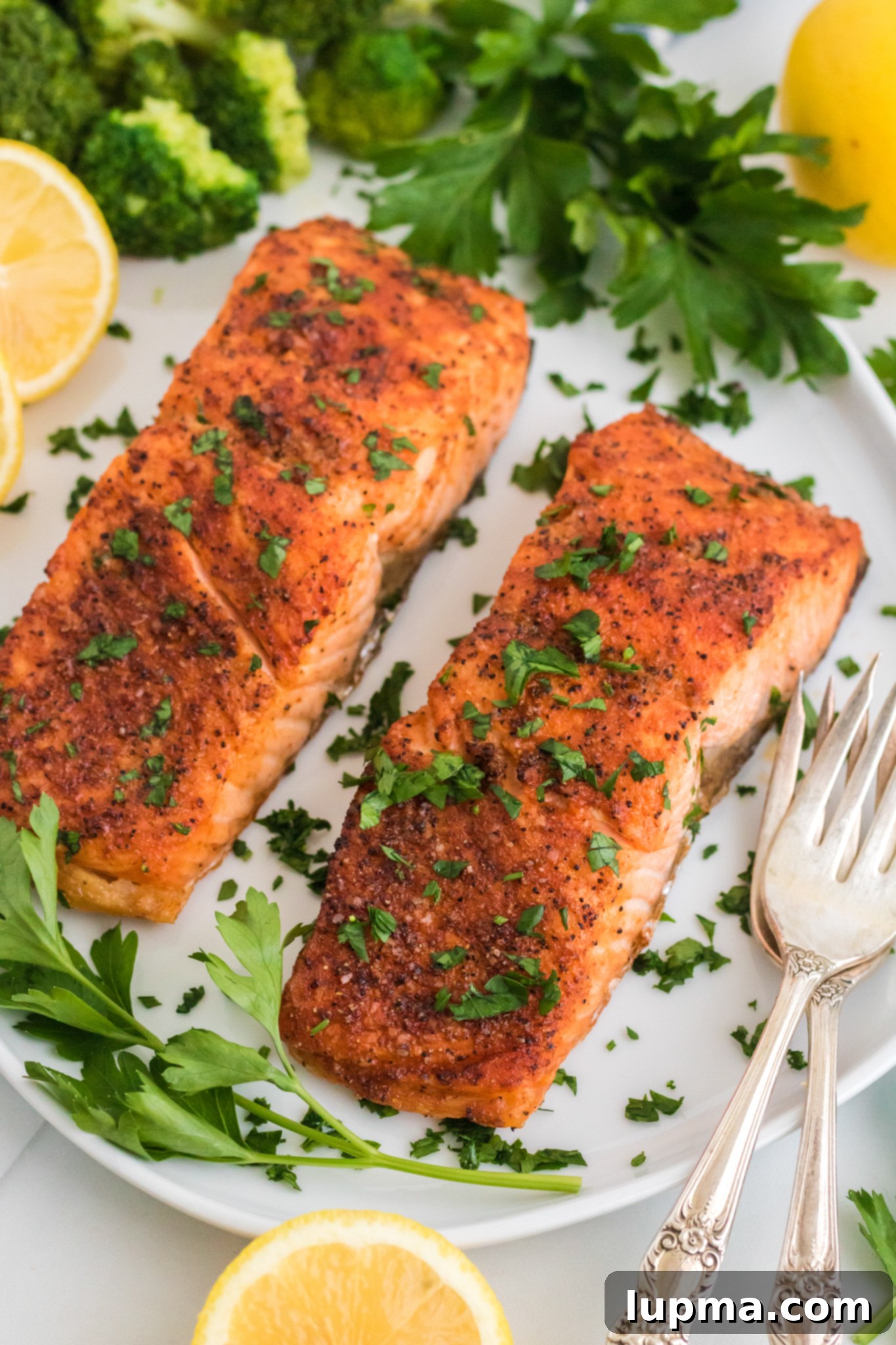 Speedy Air Fryer Salmon Perfection 2 Two perfectly seasoned and cooked salmon fillets, garnished with fresh chopped herbs, are presented on a clean white plate. The plate is beautifully arranged with vibrant sliced lemons, fresh parsley leaves, and bright green broccoli florets. Two vintage-style forks are carefully placed beside the salmon, ready for dining.