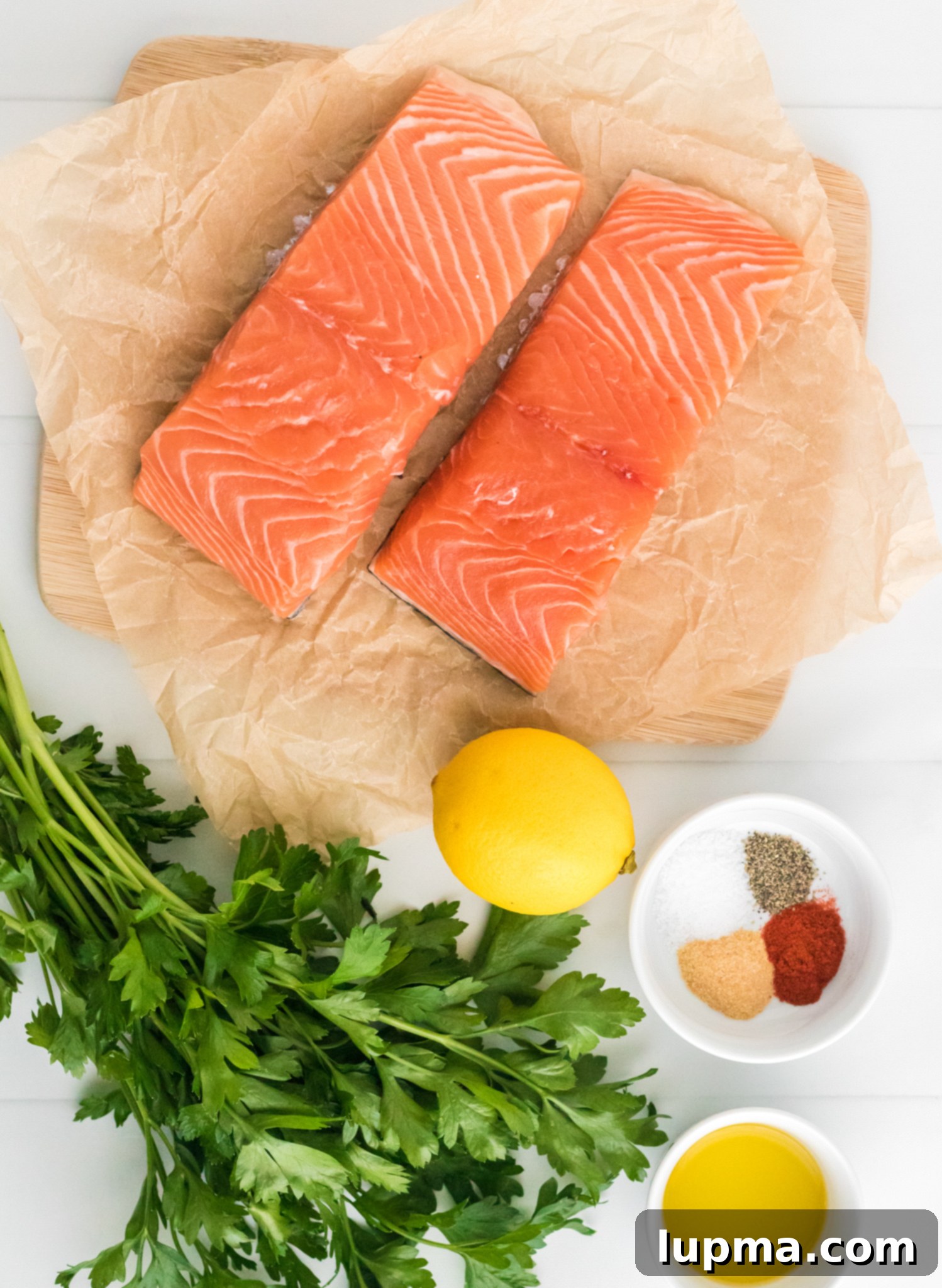 Speedy Air Fryer Salmon Perfection 3 Two fresh, raw salmon fillets are carefully arranged on parchment paper, which rests on a rustic wooden cutting board. Alongside them, a vibrant bunch of fresh parsley, a whole bright yellow lemon, and a small ceramic bowl containing a perfectly mixed blend of various spices, salt, and pepper are displayed. Adjacent to the spice bowl, another small bowl holds a glistening pool of olive oil, all set on a clean white surface, indicating preparation for a delicious meal.