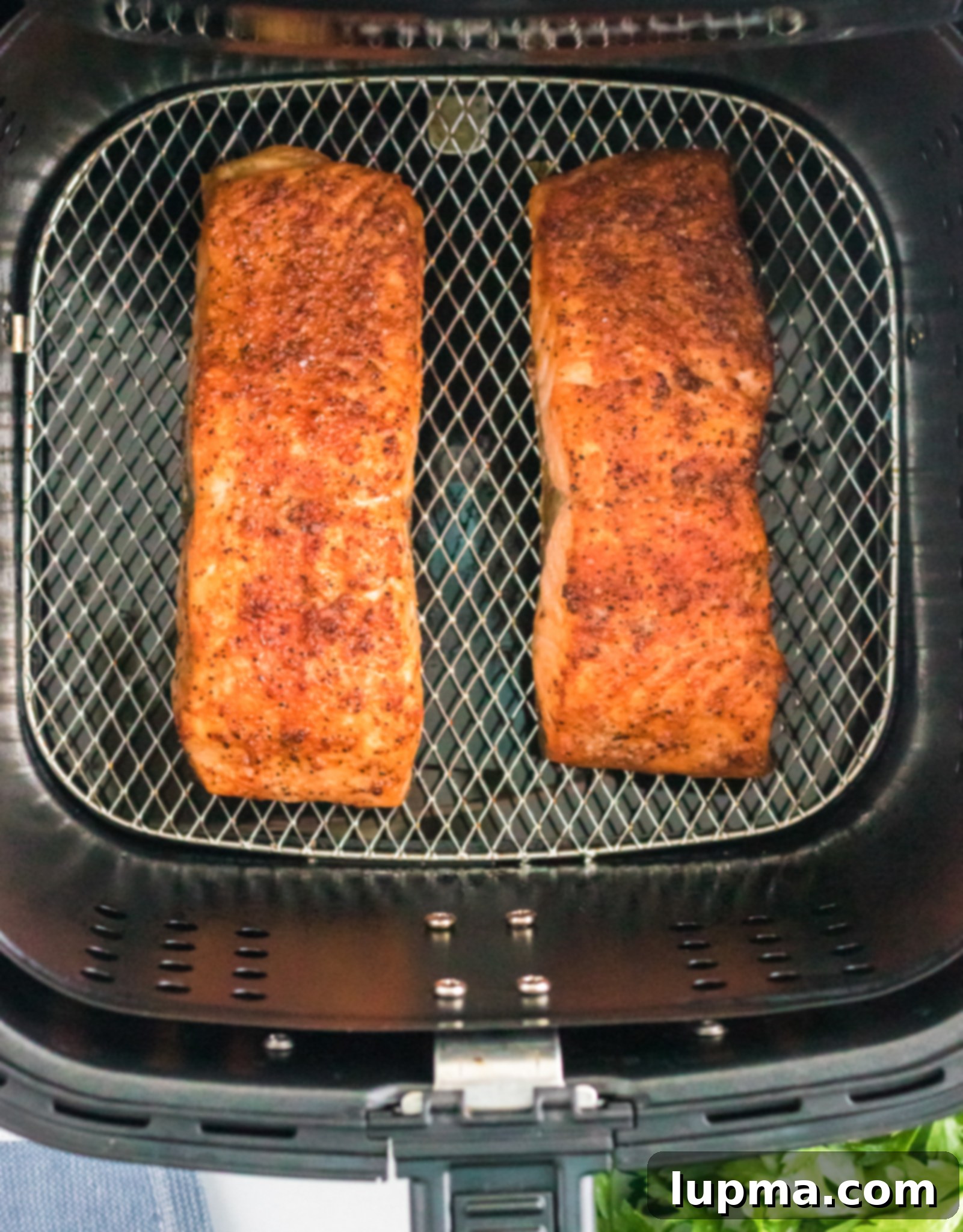Speedy Air Fryer Salmon Perfection 5 Two seasoned salmon fillets are visibly cooking inside an air fryer basket, showing a beautiful golden-brown and crispy texture. A hint of fresh green herbs is visible in the bottom right corner of the frame, suggesting a fresh garnish to come.