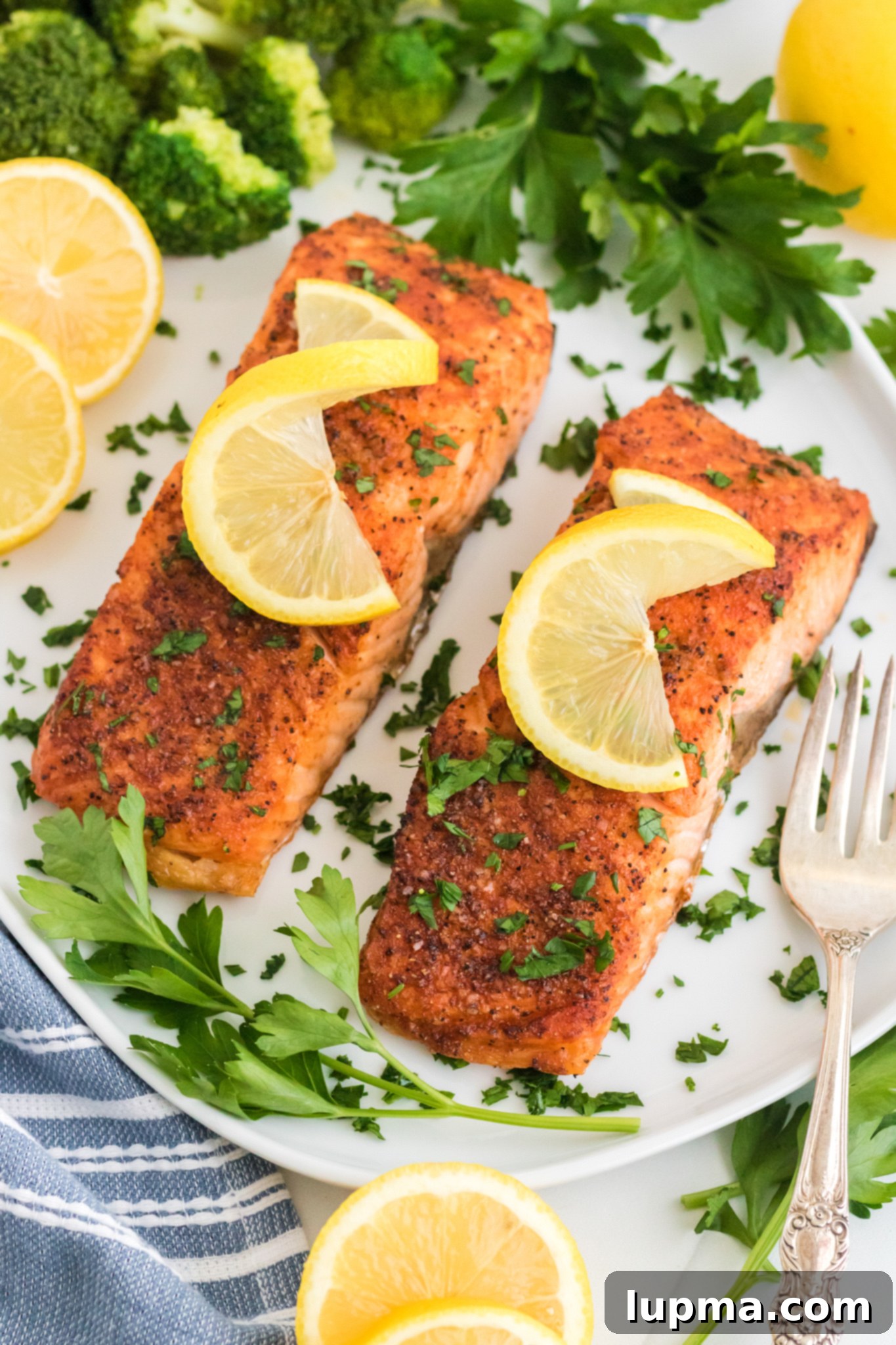 Speedy Air Fryer Salmon Perfection 6 Two perfectly cooked, seasoned salmon fillets are elegantly arranged on a white plate, each garnished with a fresh lemon slice and a sprinkle of chopped herbs. Sprigs of fresh parsley and additional lemon wedges are artfully placed around the salmon. A fork and a side of steamed broccoli are visible adjacent to the plate, completing the healthy and inviting meal.