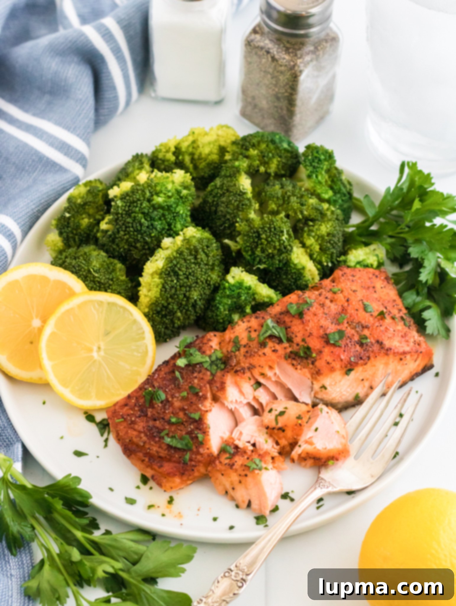 Speedy Air Fryer Salmon Perfection 7 A beautifully presented plate features roasted salmon, artfully garnished with fresh herbs, alongside vibrant steamed broccoli and bright lemon slices. A fork is gently flaking the tender salmon, showcasing its perfect doneness. In the soft-focus background, salt and pepper shakers, a refreshing glass of water, and a blue-striped cloth are visible. Nearby, a sprig of fresh parsley and a whole lemon further enhance the appealing culinary scene.