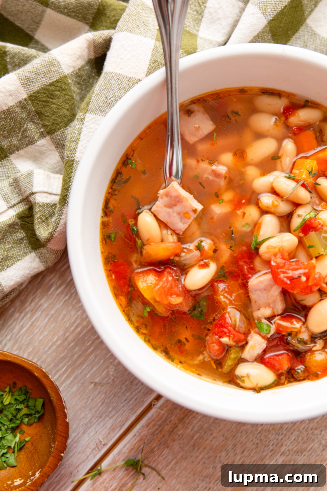 Hearty Navy Bean Soup 2 Hearty Navy Bean Soup in a white bowl with a spoon and napkin, garnished with fresh herbs.