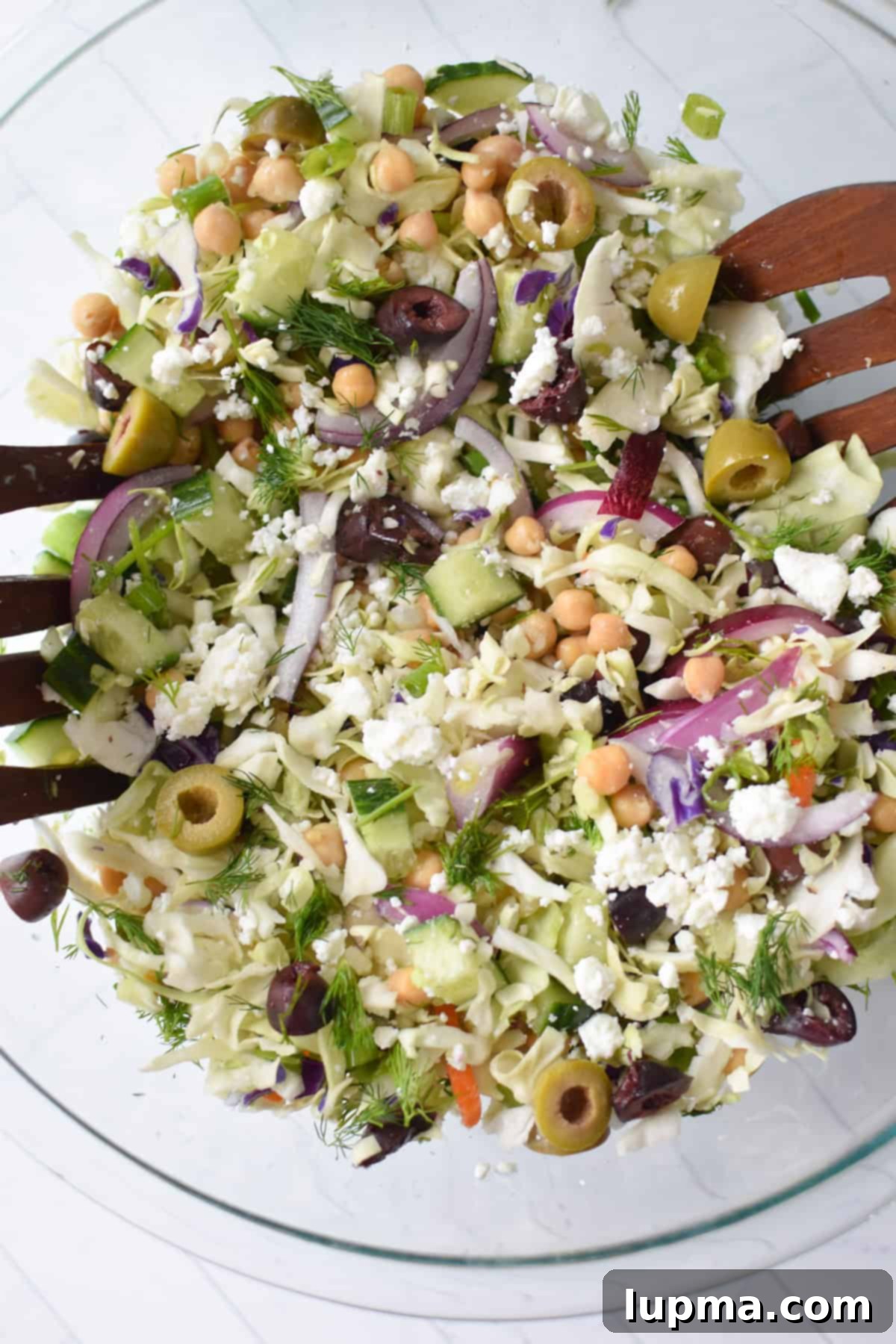 Greek Cabbage Salad in a clear glass bowl with serving spoons