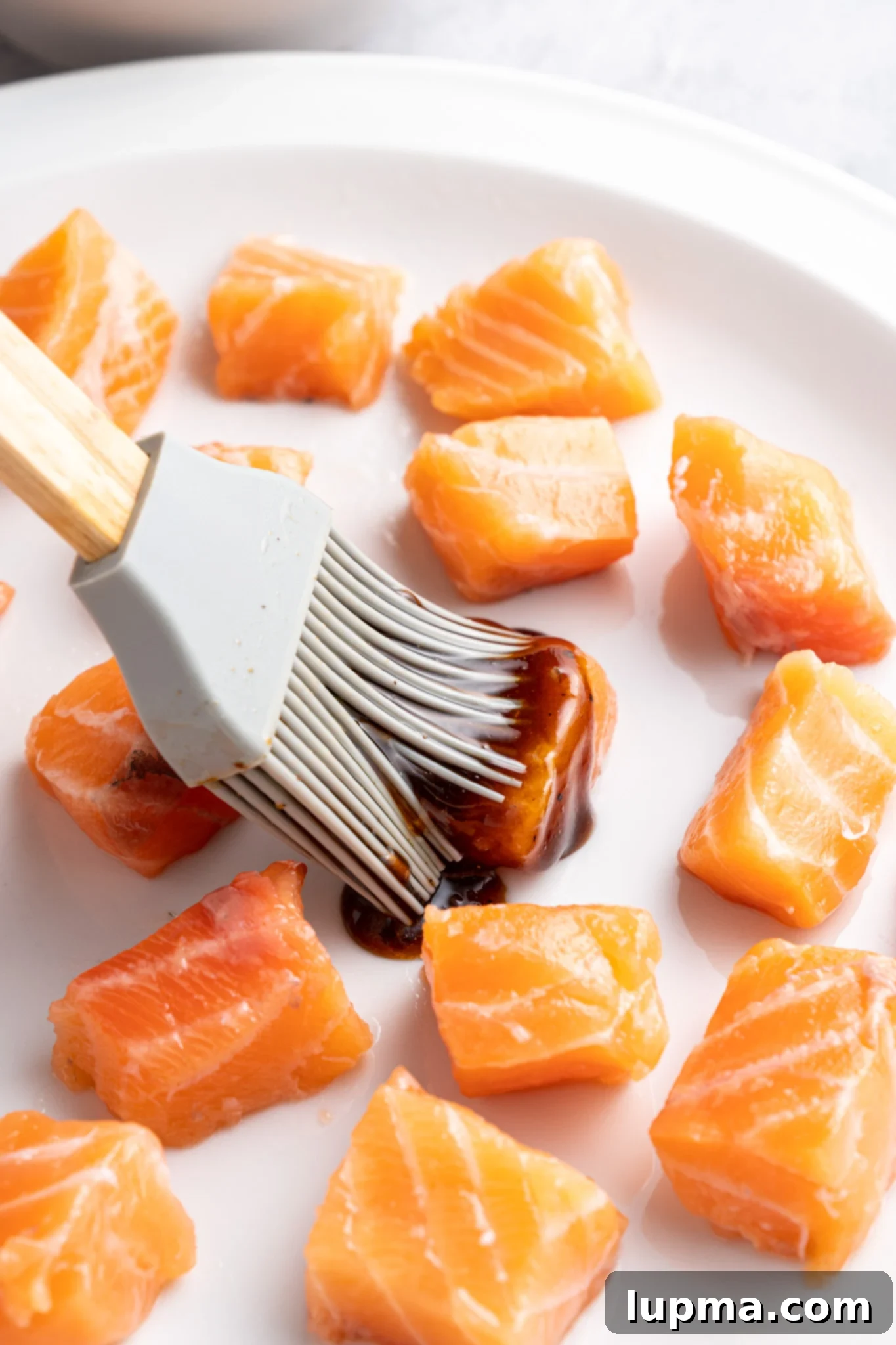 Teriyaki Salmon Bites: An Air Fryer Sensation 7 A silicone brush applies a dark marinade to raw salmon cubes on a white plate.