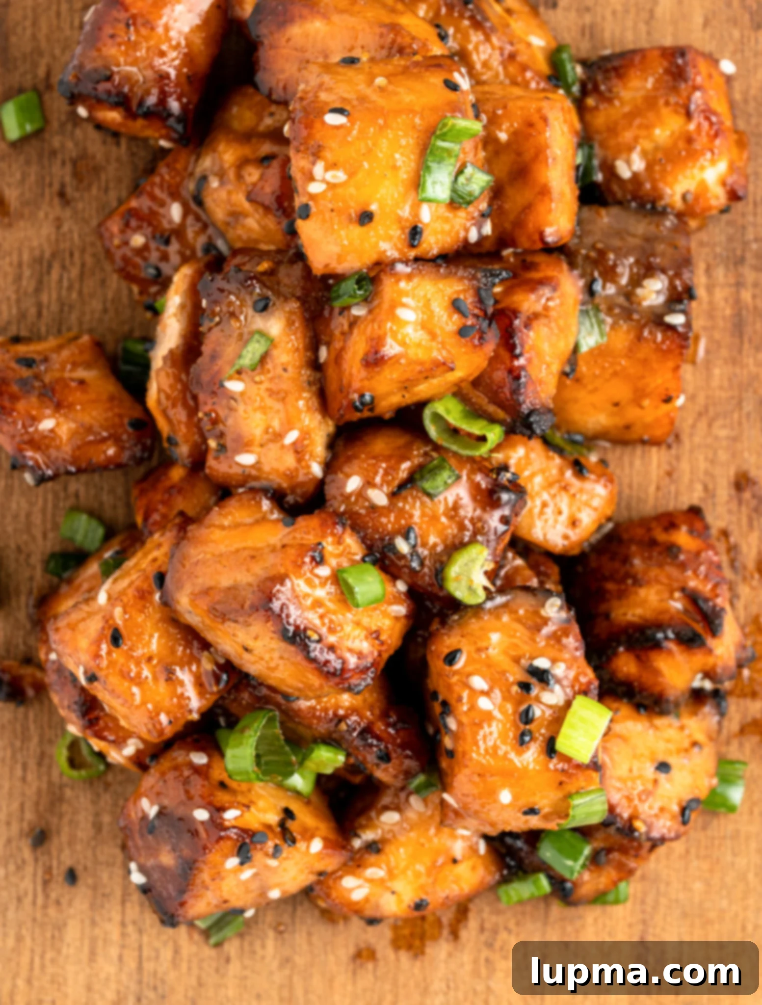 Teriyaki Salmon Bites: An Air Fryer Sensation 9 A pile of glazed, air fryer salmon bites topped with sesame seeds and sliced green onions on a wooden surface.