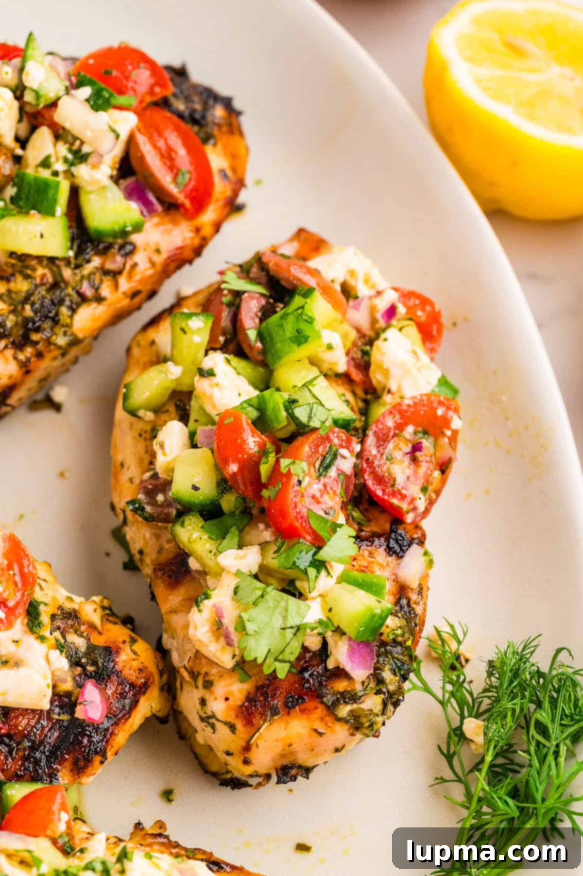 Grilled Mediterranean chicken breast topped with cucumber tomato olive and feta salad on a white plate, showcasing a healthy and delicious meal.