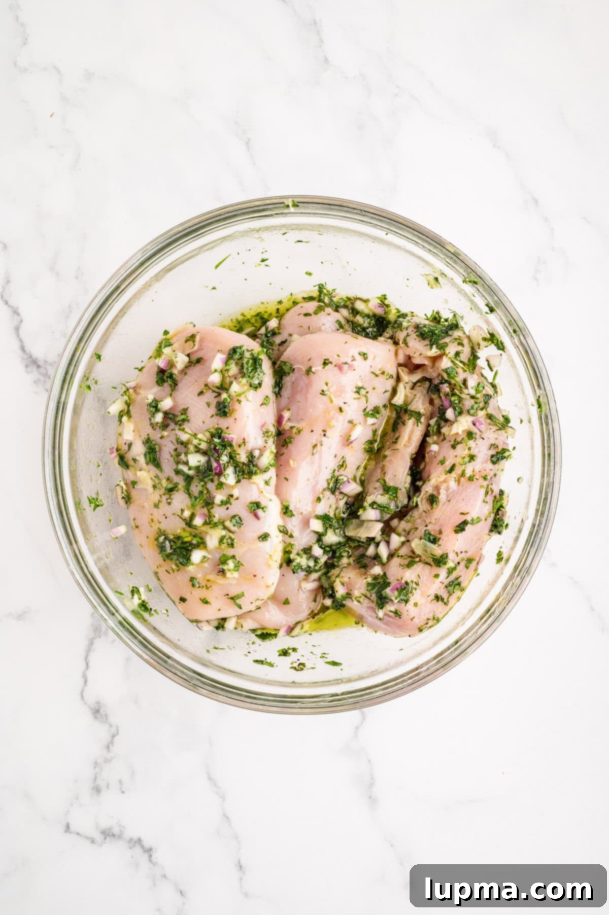 Chicken breasts marinating in a glass bowl, absorbing Mediterranean flavors.